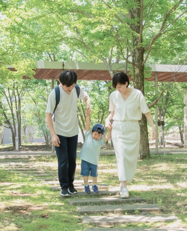 公園の木々に囲まれた遊歩道で、両親が幼い子供の手を引いて歩いている様子。家族の温かい絆を感じさせる風景。