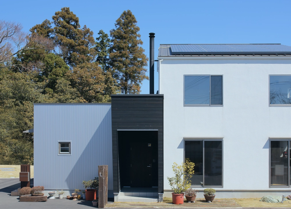 白い外壁と黒い玄関が特徴的なモダンな住宅。周囲には植栽と自然が広がる。